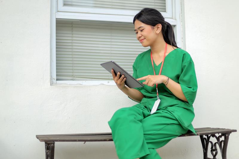 smiling-asian-woman-wear-nurse-uniform-using-digital-tablet-while-sitting-bench
